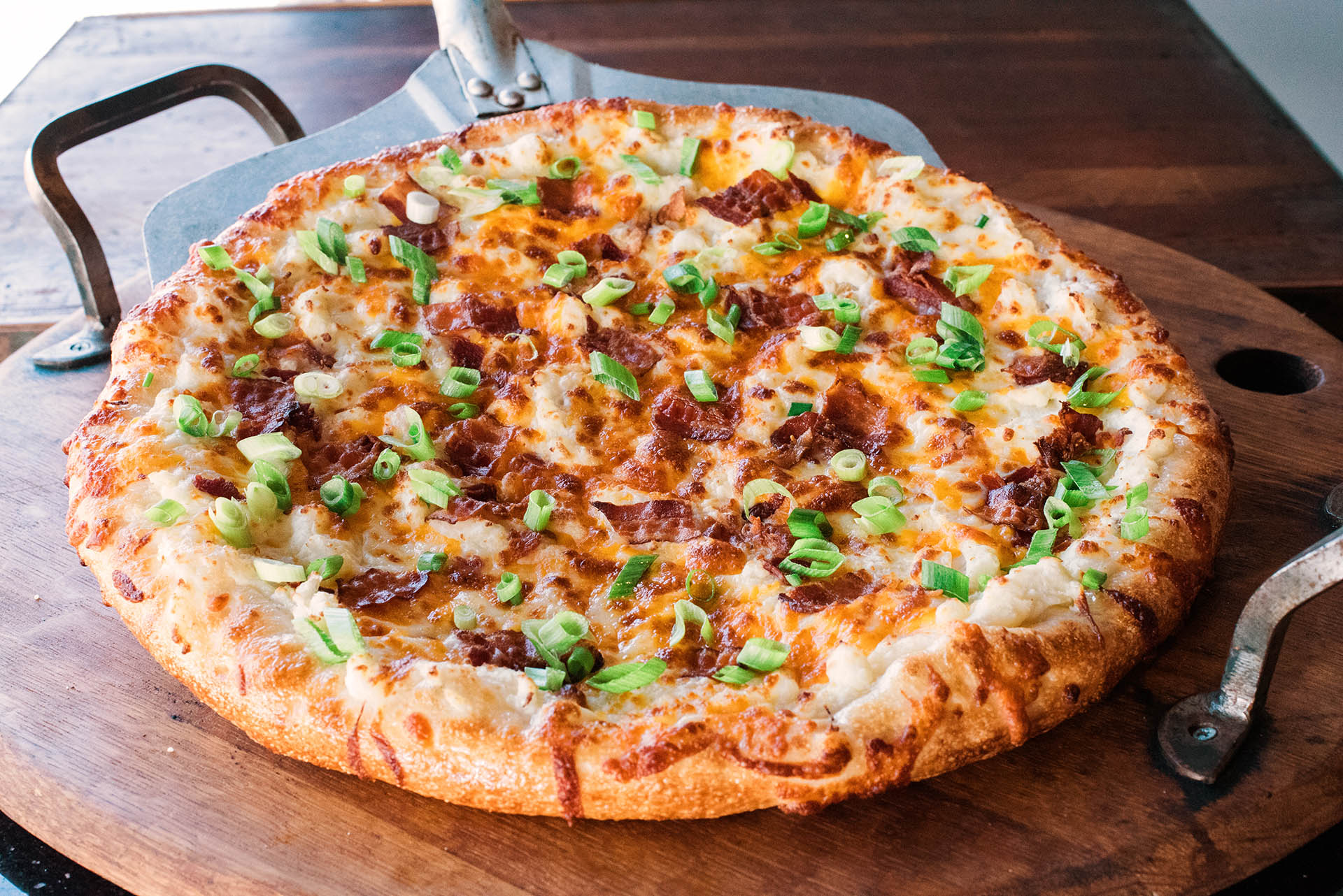 A mashed potato pizza with chives, cheddar and bacon presented on a rustic wooden serving platter.